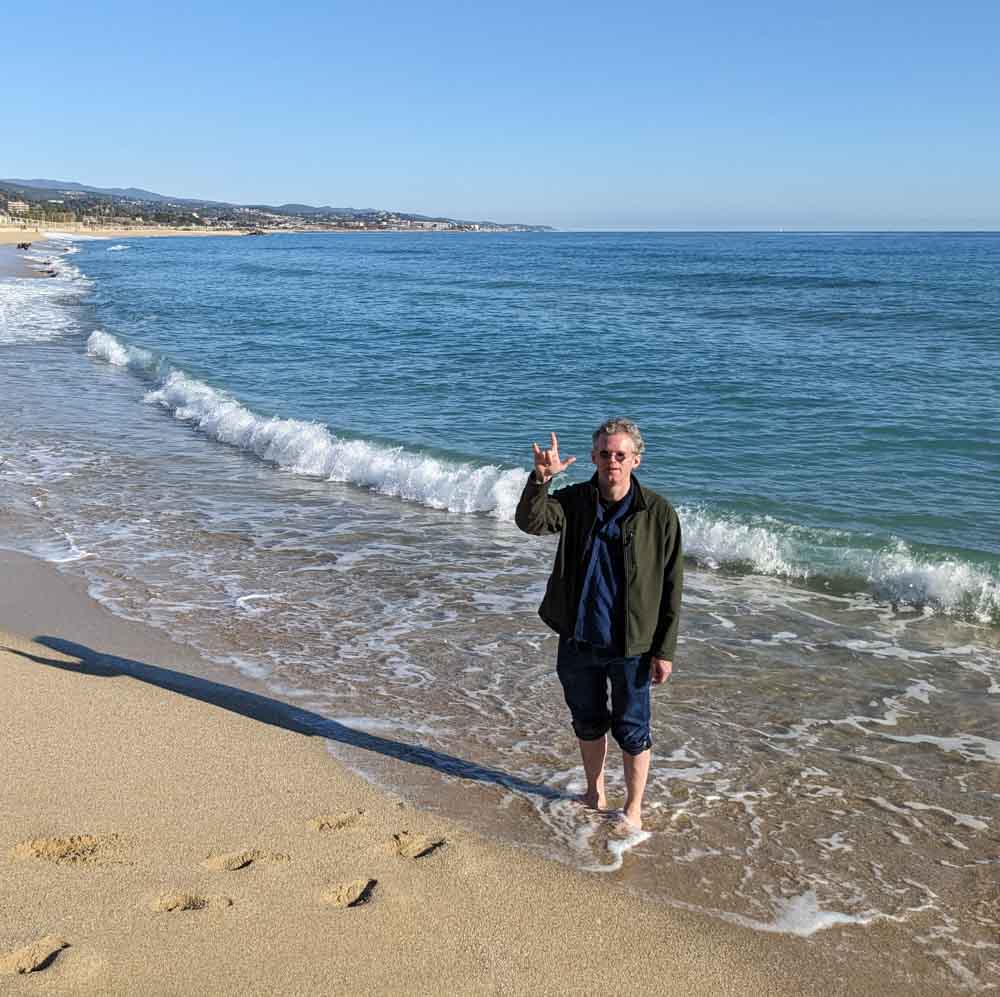 Jonas Hauer steht barfuß am Sandstrand. Das Meer bildet sanfte Wellen und Fussabdrücke im Sand sind zu sehen. Er hat die Hand gehoben, Daumen, Zeigefinger und kleiner Finger sind abgespreizt - das Zeichen aus der amerikanischen Gebärdensprache für I love you.